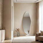 Aurora irregular curve mirror leaning against living room wall with vase, sofa and curtain in modern interior.