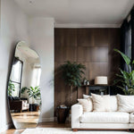 Luna irregular mirror leaning in living room with white sofa, dark wood wall and potted plants reflecting window light.