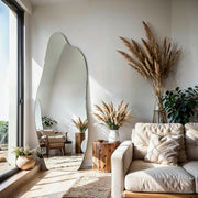 Luna irregular mirror leaning against wall in sunlit living room with sofa and pampas grass.