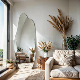 Luna irregular mirror leaning against wall in sunlit living room with sofa and pampas grass.