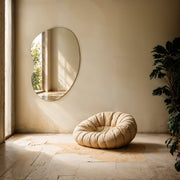 Miroir organique sans cadre Orta sur un mur enduit baigné de soleil, à côté d’un pouf moelleux