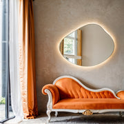 Miroir ovale sans cadre Piano sur un mur enduit texturé, au-dessus d’une méridienne en velours orange, présenté avec une lumière chaude derrière