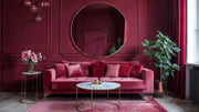 Rose gold round mirror hanging on deep red wall above pink velvet sofa, marble coffee table and potted plants by window.