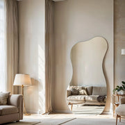 Serene irregular curve frameless mirror leaning against tall beige curtains in living room with beige sofa, floor lamp and potted plant.