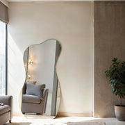 Serene curve irregular frameless mirror leaning against wall in bright living room with potted tree and grey armchair.