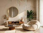 Solaro round mirror with brushed gold frame above wooden console and beige armchair in modern living room interior.